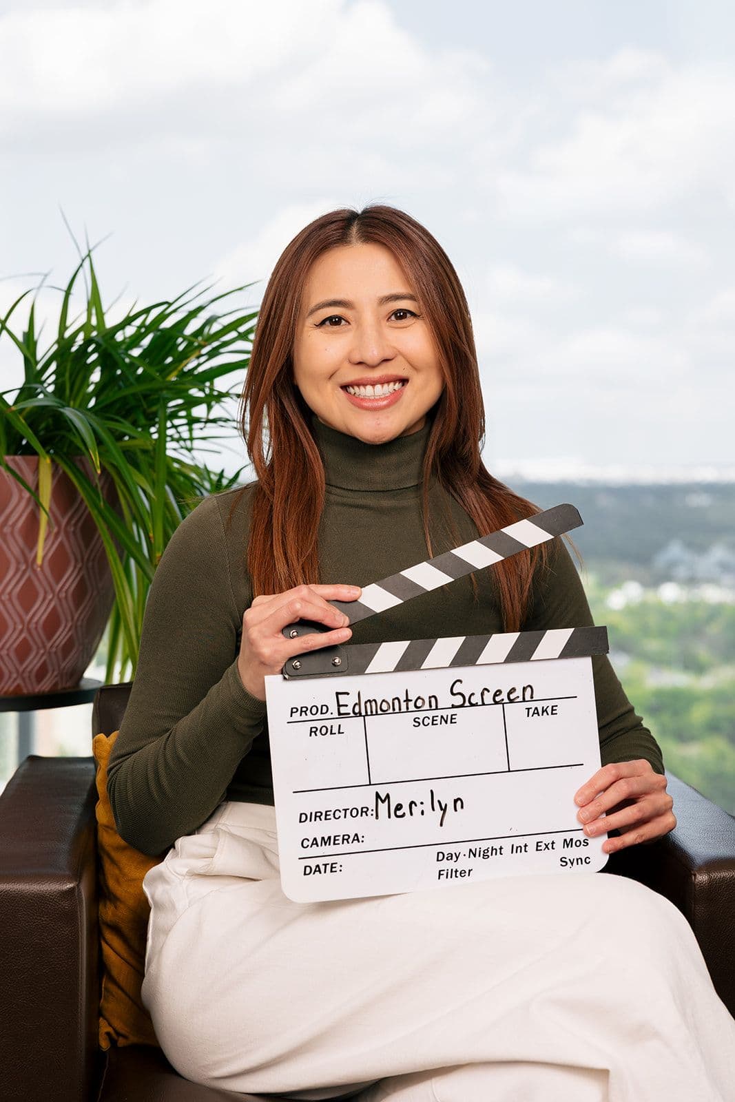 Edmonton Screen Communications and Social Media Consultant, Merilyn Tuazon, seated in front of a window holding an Edmonton Screen clapperboard.