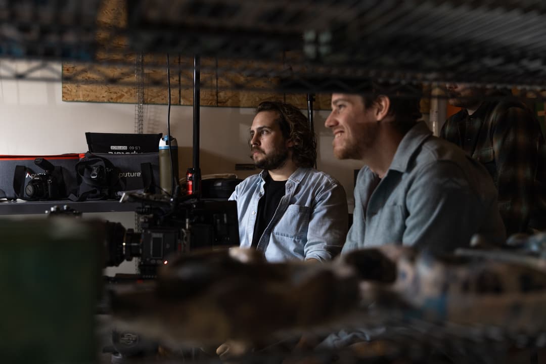Two filmmakers, including Colin Waugh, sit beside a professional camera setup inside a studio, monitoring a shoot behind shelving and equipment during production.