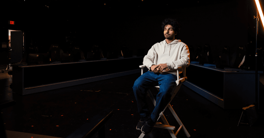 Nazzal Naseer sits in a white director’s chair in a dark studio, with rows of gaming desks and chairs behind him.