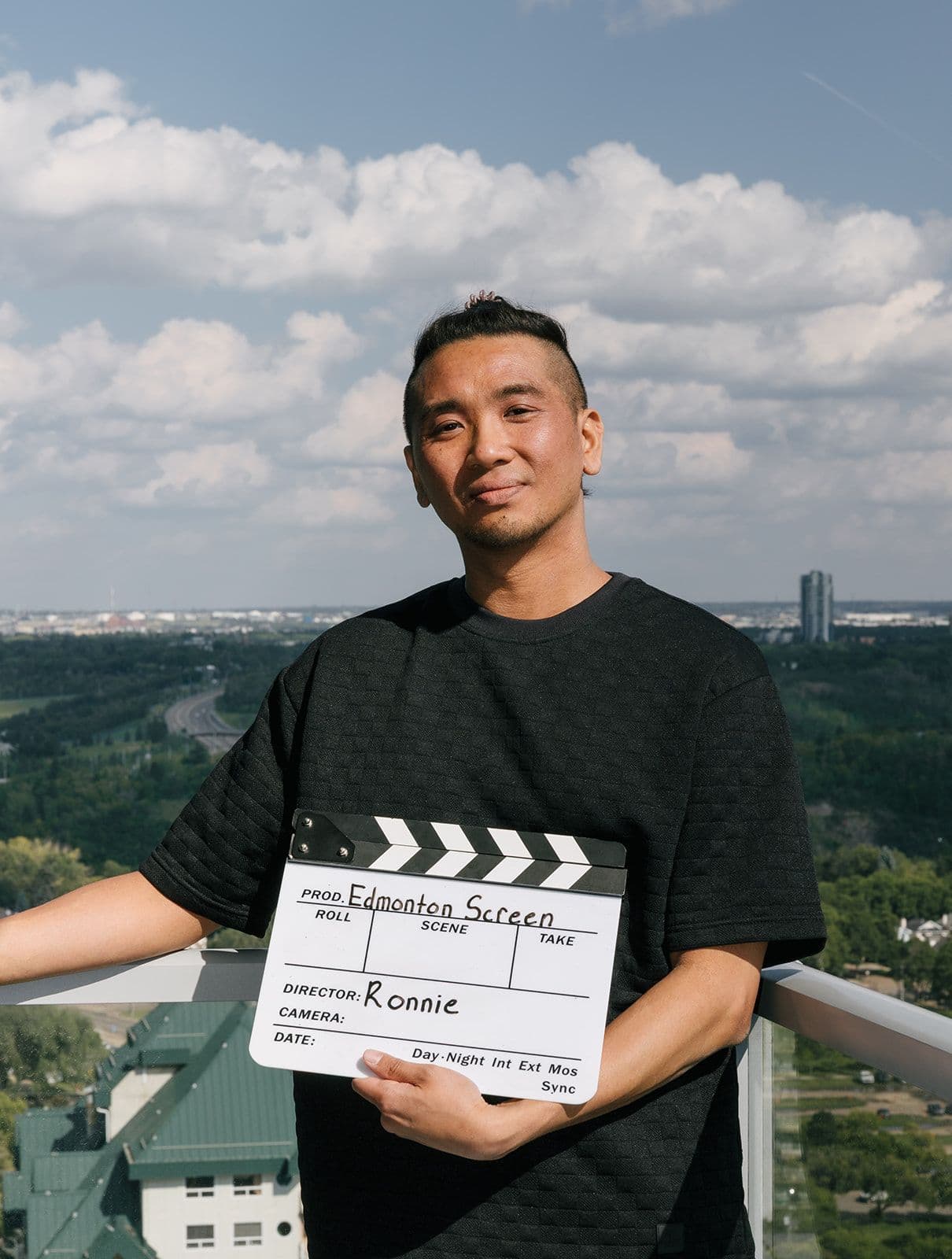 Ronnie Villanueva, Digital Media and Technology Specialist at Edmonton Screen, holding an Edmonton Screen clapperboard on a balcony overlooking the city.