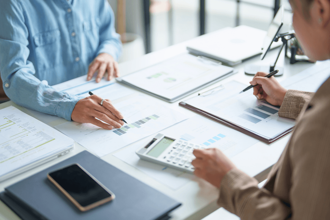 Two finance professionals reviewing bar charts and financial reports at a desk, with a calculator, pen, and smartphone visible."